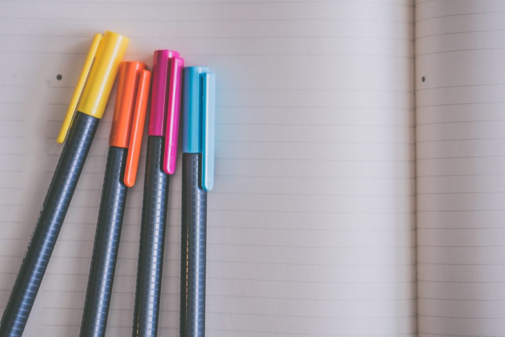 Open ruled notebook with a yellow, orange, pink and blue pens on it.