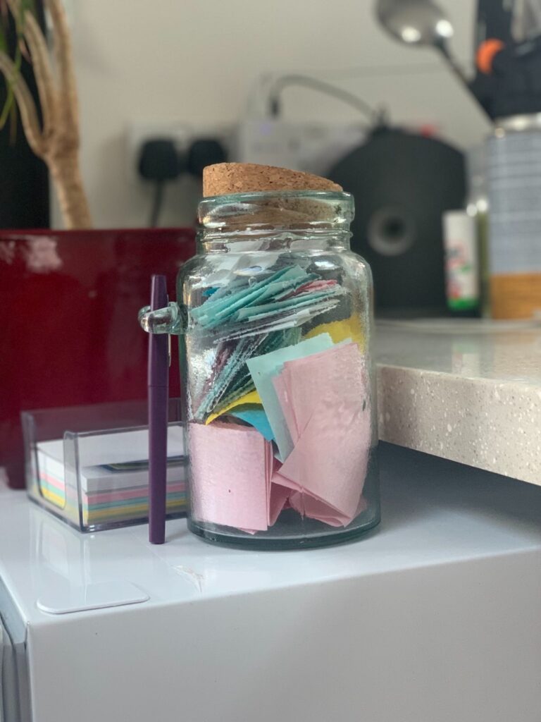 Clear green jar with a cork stopper filled with coloured notes next to a retro brown plastic cube that holds little squares of coloured paper.