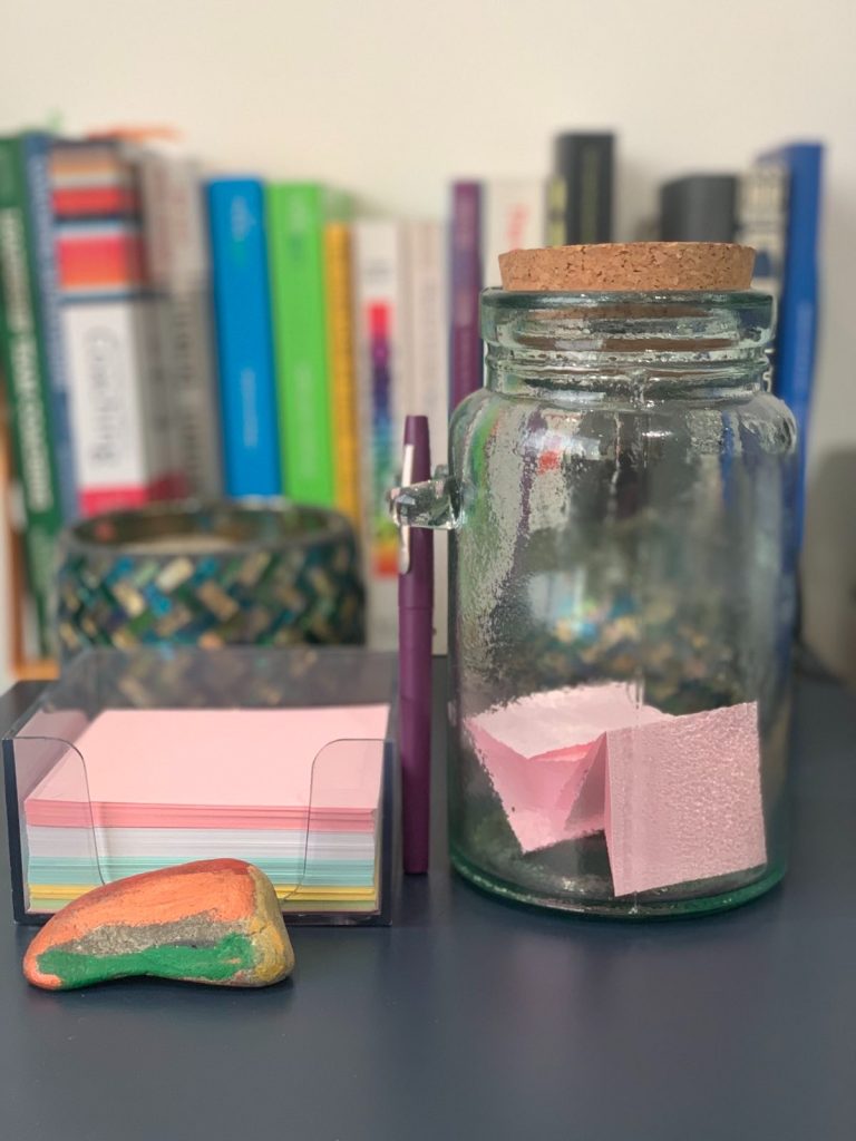 A picture of my joy jar. On the left of the picture is a plastic cube holding coloured paper with a painted rock in front of it. Next to the cube is glass jar with a cork stopper filled with some folded pieces of paper. There are books in the background.