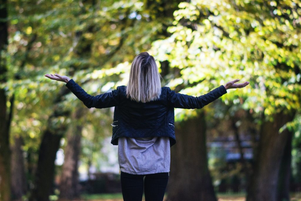 A person stands in the centre of the picture with their back to us, holding their arms up in a why gesture. The background is blurred trees.