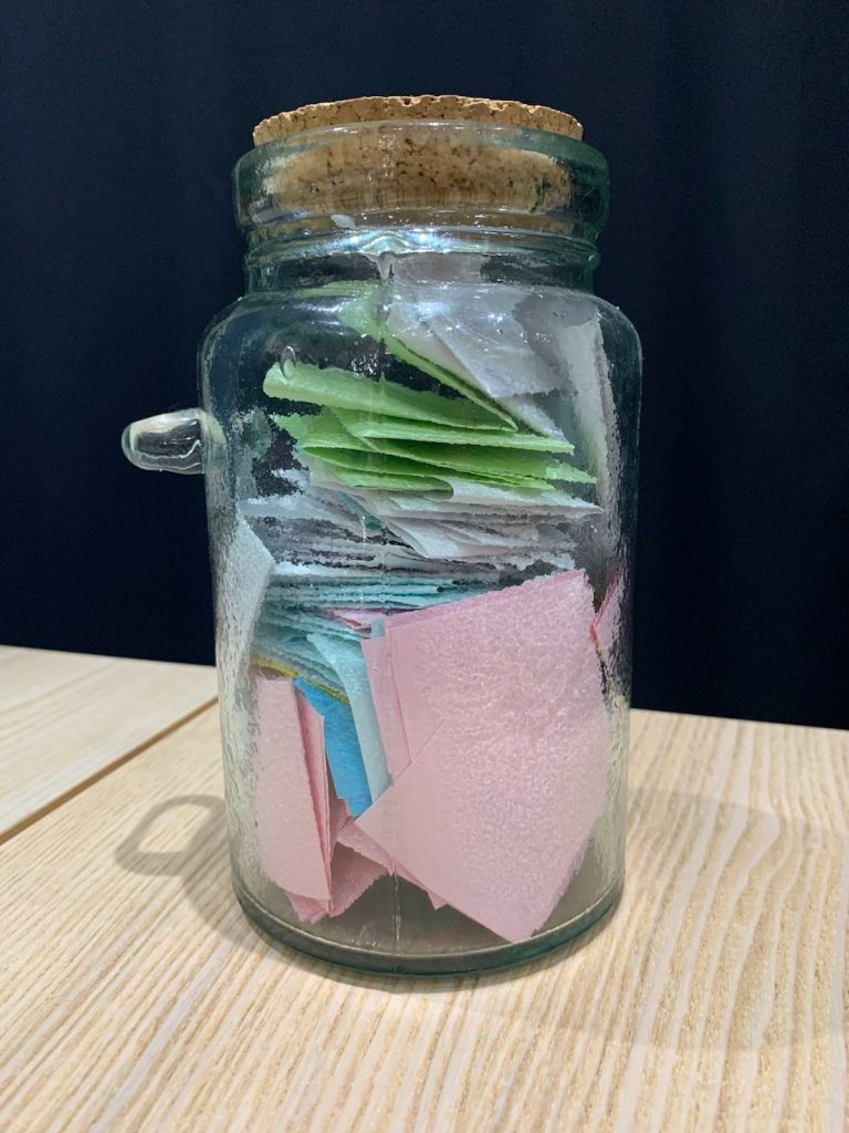 A clear glass jar with a cork stopper containing lots of coloured notes.